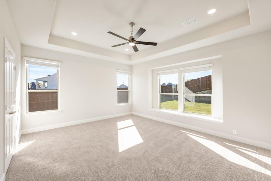 Empty bedroom with tray ceiling, ceiling fan, carpet flooring, and multiple windows with backyard views Empty bedroom with tray ceiling, ceiling fan, carpet flooring, and multiple windows with backyard views
