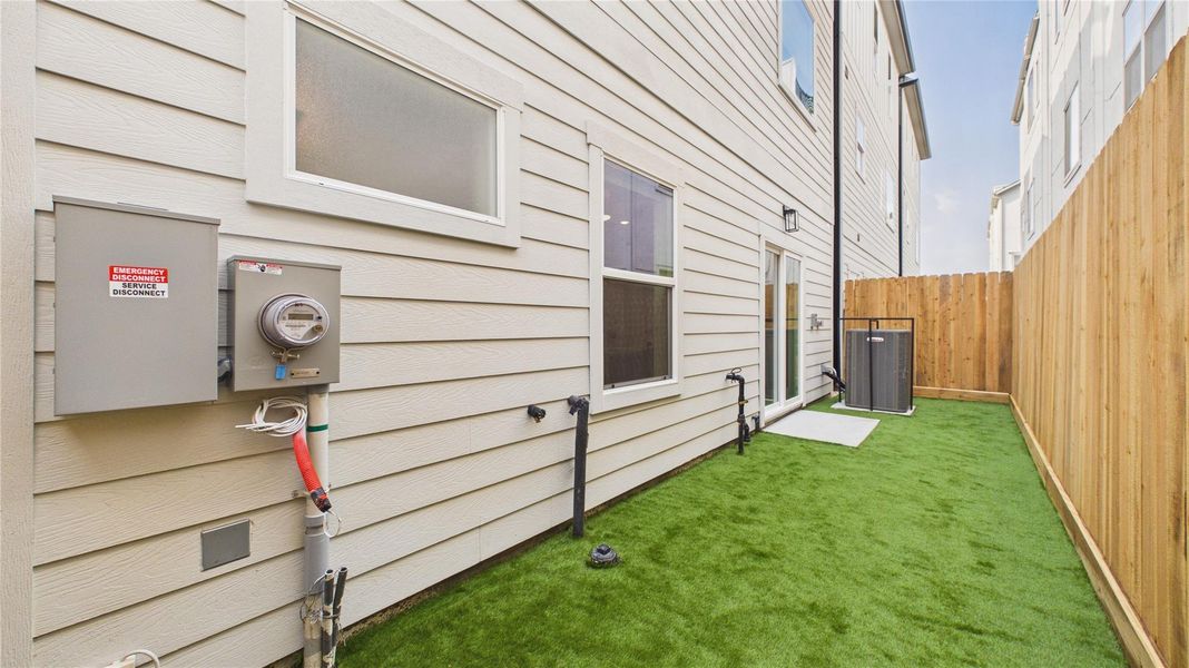 This photo shows a narrow side yard with artificial grass, bordered by a wooden fence. It features an exterior wall with a utility meter and a small patio area near a glass door.