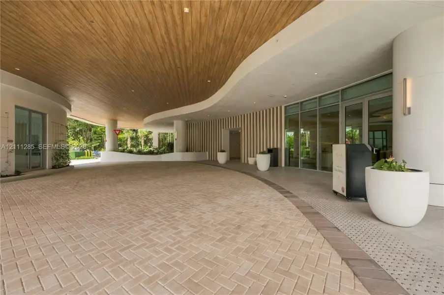 Covered porte-cochère featuring a curved wood-panel ceiling, herringbone-pattern paver drive, vertical slat wall detailing, and expansive glass entry doors Covered porte-cochère featuring a curved wood-panel ceiling, herringbone-pattern paver drive, vertical slat wall detailing, and expansive glass entry doors