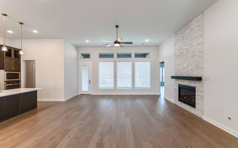Open-concept living area with wood-finish flooring, a contemporary fireplace featuring a light stacked stone surround and dark mantel, and multiple windows Open-concept living area with wood-finish flooring, a contemporary fireplace featuring a light stacked stone surround and dark mantel, and multiple windows