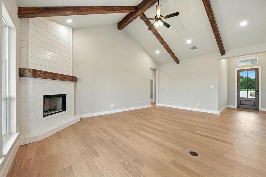 Unfurnished living room with a fireplace, beam ceiling, ceiling fan, light wood finished floors, and high vaulted ceiling Unfurnished living room with a fireplace, beam ceiling, ceiling fan, light wood finished floors, and high vaulted ceiling