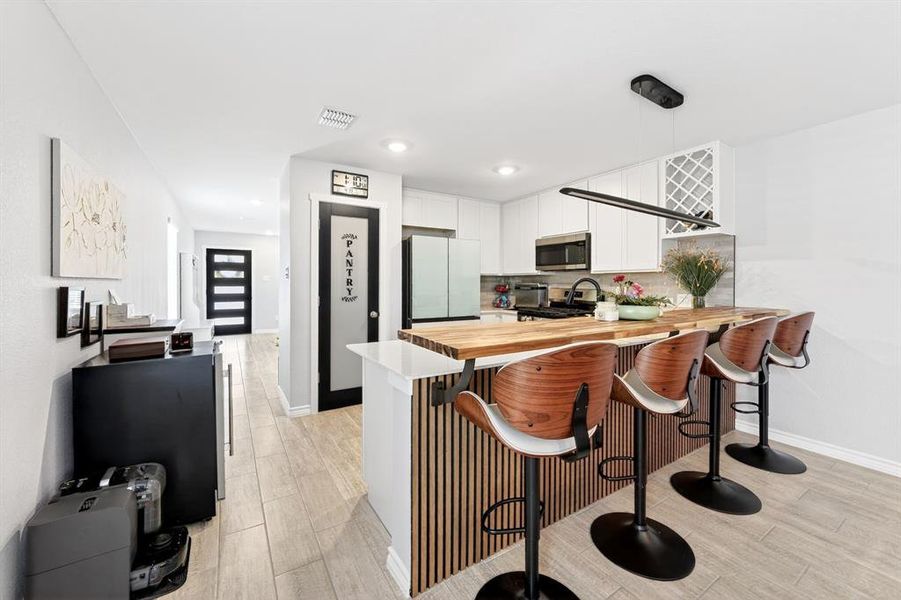 Modern kitchen featuring an extended butcher block countertop with breakfast bar seating, white shaker cabinetry, stainless steel appliances, and a built-in wine rack
