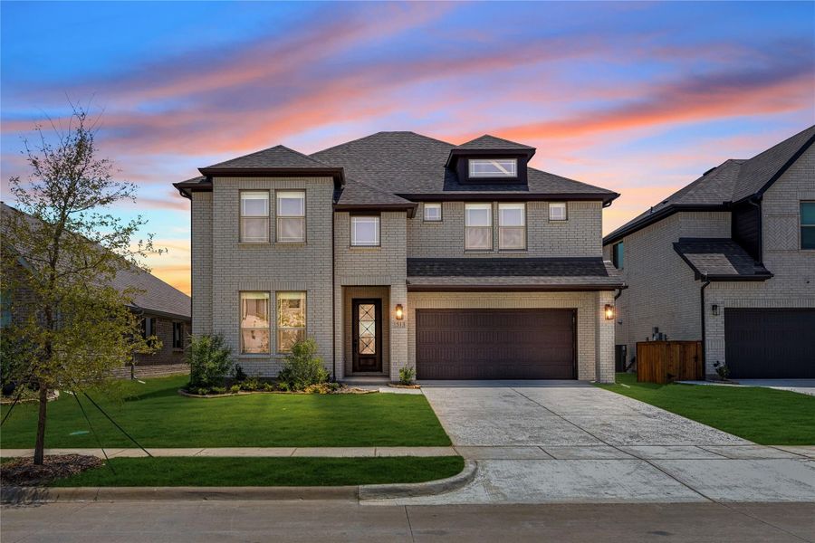 Two-story brick home with two-car garage, dark roof, and manicured front lawn at dusk