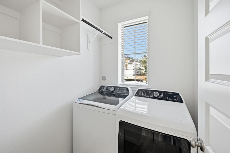 Bright and functional laundry room featuring modern washer and dryer units, ample shelving, and a window for natural light.