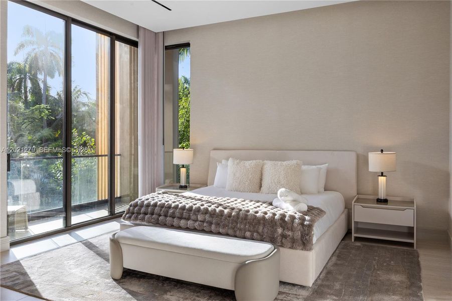 Clean-lined en-suite bedroom with upholstered king bed, matching nightstands, neutral textured walls, area rug, light stone floors, sheer drapery, and corner floor-to-ceiling sliding glass doors opening to glass-railed balcony with tropical garden views.