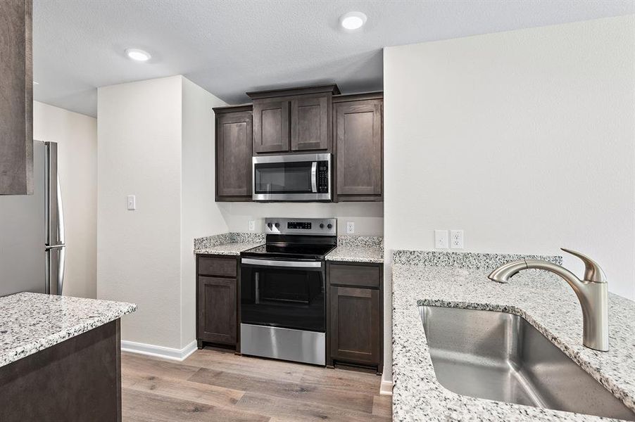 Kitchen with stainless steel appliances, dark brown cabinets, light wood-style floors, light stone countertops, and recessed lighting