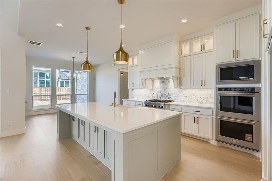 Kitchen with glass insert cabinets, appliances with stainless steel finishes, tasteful backsplash, a kitchen island with sink, and hanging light fixtures