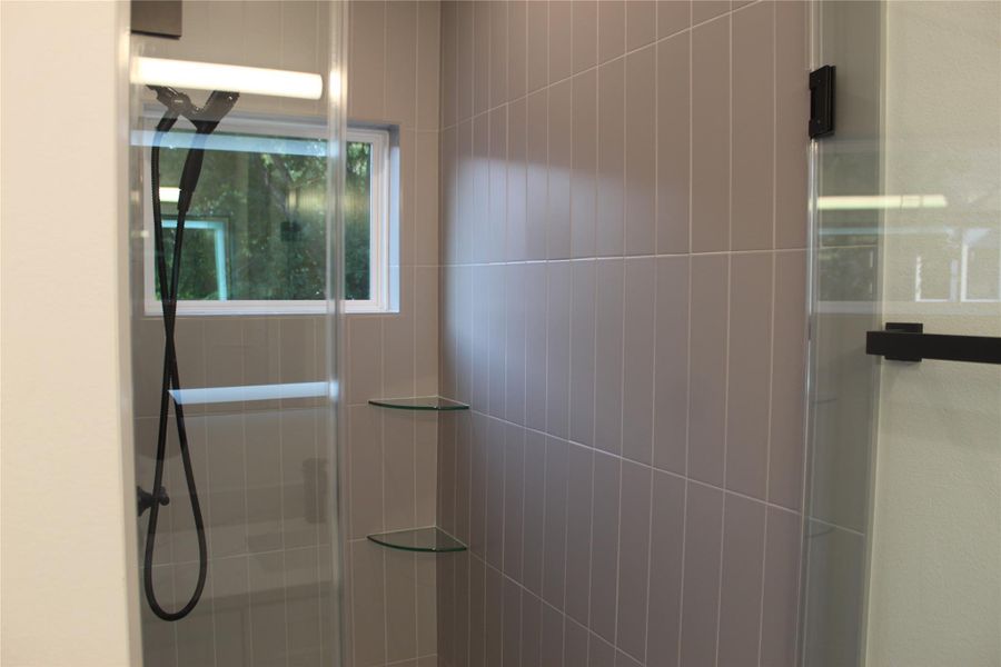 A well-appointed shower space offers sleek tile work, clear glass shelving, and a view of verdant surroundings.