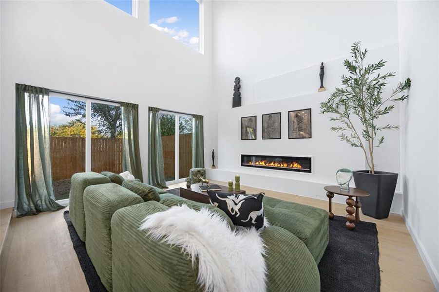 Living room featuring plenty of natural light, light wood-style floors, a high ceiling, and a glass covered fireplace