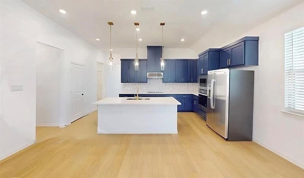 Kitchen with appliances with stainless steel finishes, a kitchen island with sink, blue cabinetry, hanging light fixtures, and light wood-style floors