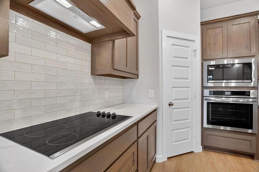Kitchen featuring light wood-finish cabinetry, white subway tile backsplash, white countertops, a built-in cooktop, and stainless steel double oven