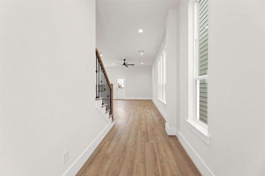 Bright hallway with wood like flooring, recessed lighting, and a ceiling fan. Large windows on one side enhance the natural light. A staircase with a wooden banister leads to the upper level.
