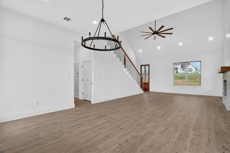 Open-concept living area featuring wood-finish flooring, a contemporary chandelier, a vaulted ceiling with a ceiling fan, a staircase with wood handrails and black balusters, and a large window