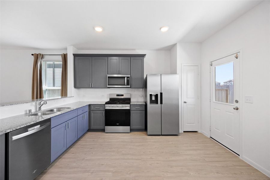 Modern kitchen with sleek gray and blue cabinetry, stainless steel appliances, and a dual sink. Features a bright, open layout with wood-look flooring and a door leading outside.