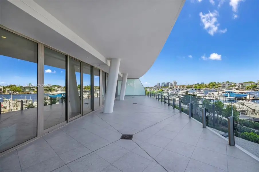 Expansive covered terrace featuring large-format tile flooring, structural support columns, floor-to-ceiling glass wall panels, and a frameless glass railing