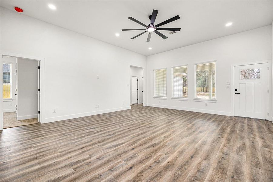 This spacious living area features modern wood flooring, high ceilings with a stylish ceiling fan, and abundant natural light through large windows. The neutral color palette and open layout offer a versatile space for personalization.