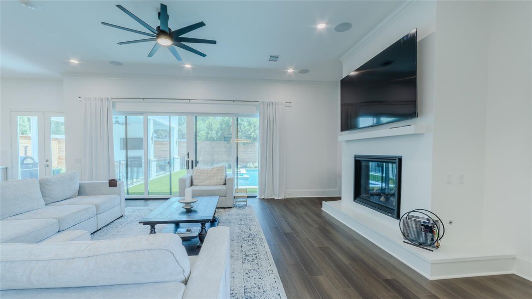 This spacious, modern living room features a large sectional sofa, sleek ceiling fan, and a mounted TV above a fireplace. Glass sliding doors open to a view of the pool and backyard, inviting ample natural light.