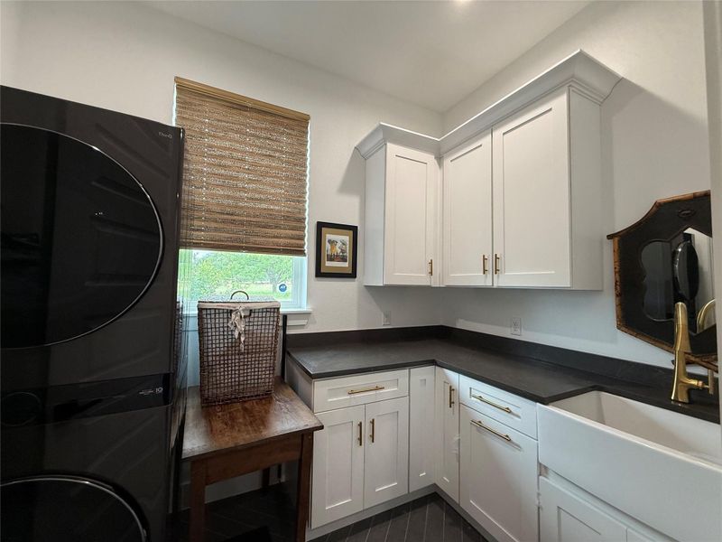 Laundry room featuring white shaker cabinetry, dark countertops, and a white farmhouse sink with a gold-tone faucet