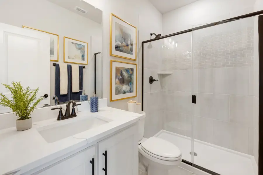 Bathroom with white vanity, glass-enclosed tile shower, toilet, and framed wall art with dark hardware accents. Bathroom with white vanity, glass-enclosed tile shower, toilet, and framed wall art with dark hardware accents.