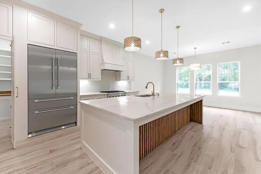 This modern kitchen features a spacious island with a marble countertop and wood accents, pendant lighting, and a large stainless steel refrigerator. The open layout allows for natural light from the large windows, creating a bright and inviting space.