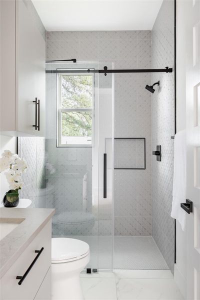 The bathroom features a tiled shower with black water fixtures and a built-in cubby, while ample natural light enhances the modern, spa-like ambiance.