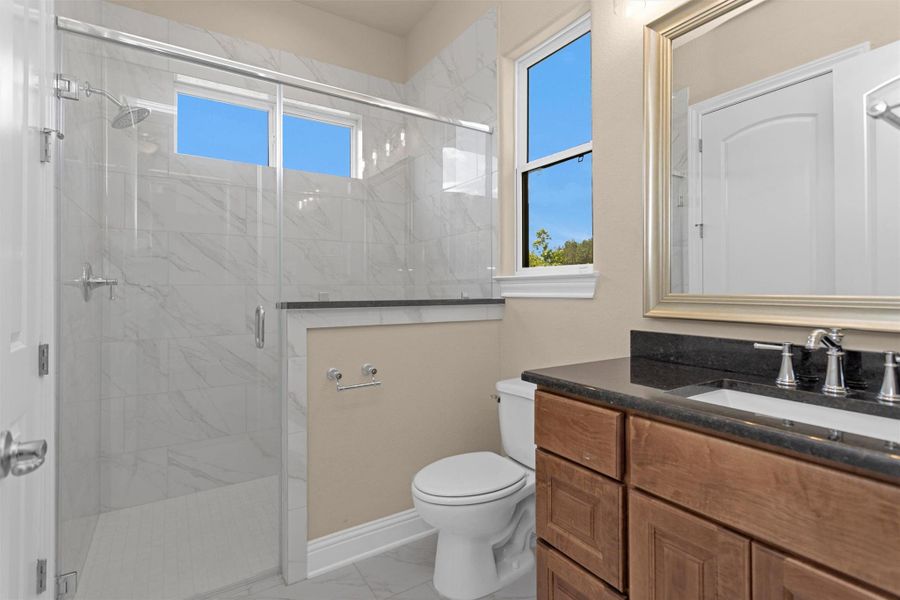 This en-suite bathroom features a spacious walk-in shower with elegant marble-style tile, natural light from dual windows, and warm wood cabinetry topped with sleek black granite counter. This en-suite bathroom features a spacious walk-in shower with elegant marble-style tile, natural light from dual windows, and warm wood cabinetry topped with sleek black granite counter.