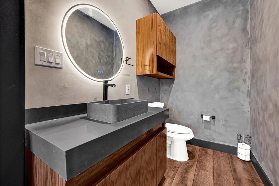 Sleek grey countertop with a rectangular vessel sink, a round illuminated mirror, and stylish wood cabinetry. The textured grey walls and wooden flooring add a contemporary touch, while the toilet area is neatly arranged.