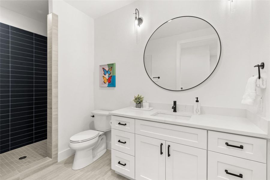 This bathroom features a modern design with a spacious walk-in shower, sleek black tiles, and a white vanity with black hardware. It includes a large round mirror, contemporary lighting, and a minimalistic aesthetic.