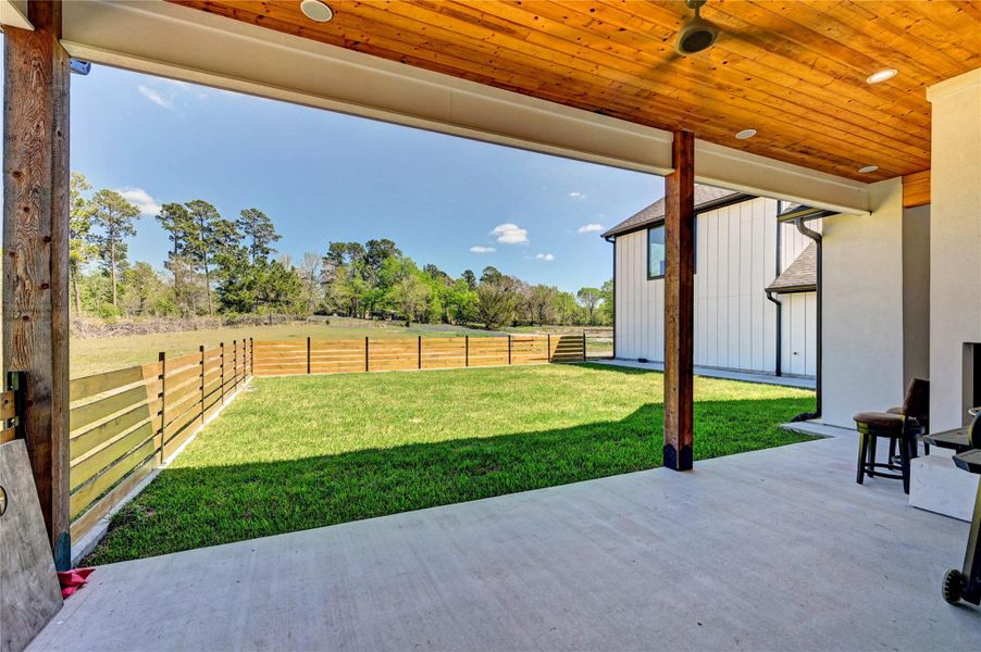 This photo showcases a spacious covered patio with a wooden ceiling, overlooking a large, fenced backyard with lush green grass. The area offers a serene view of surrounding trees, ideal for outdoor relaxation and entertainment.