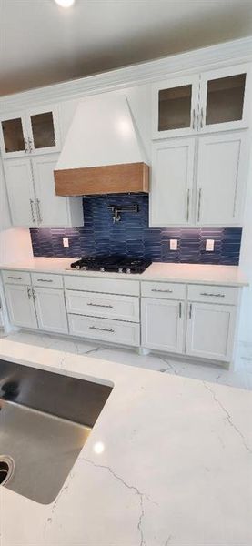 Kitchen with light stone counters, tasteful backsplash, white cabinetry, custom range hood, and gas stovetop