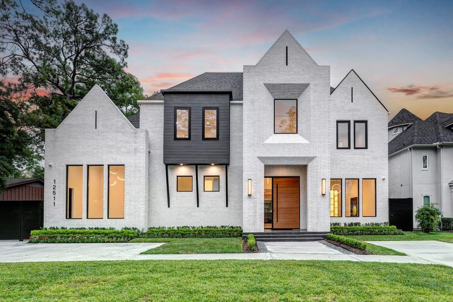 This modern, two-story home features a striking white brick exterior with black accents and large windows. The front yard is landscaped with neatly trimmed shrubs and a wide driveway. The architecture is contemporary with clean lines and a welcoming entrance.