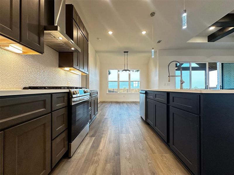 Kitchen featuring light wood finished floors, stainless steel appliances, backsplash, wall chimney range hood, and decorative light fixtures