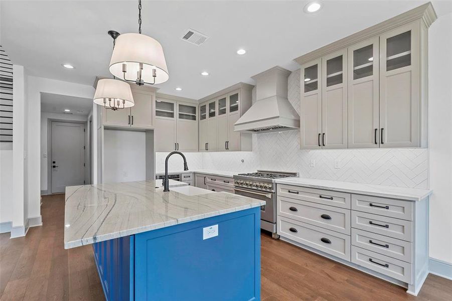 Kitchen with stainless steel stove, decorative light fixtures, light stone countertops, custom range hood, and a center island with sink