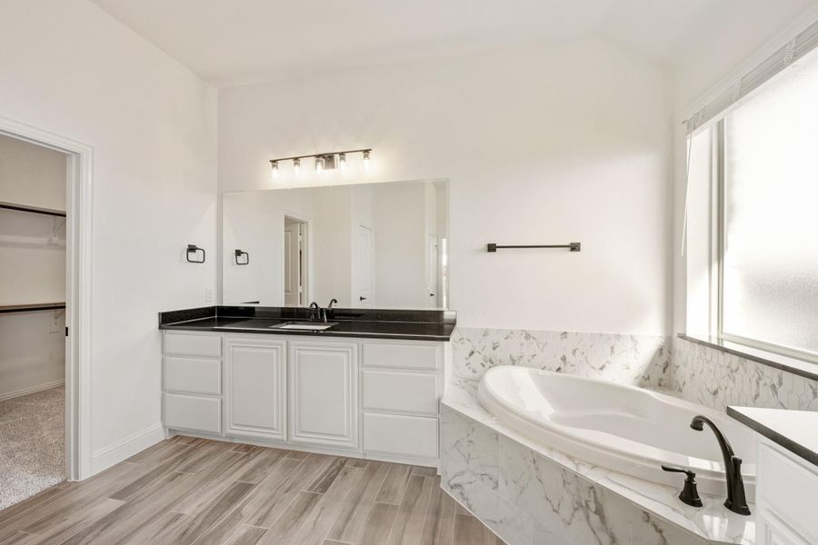 Master bathroom with white cabinets, dark countertop, marble soaking tub, and wood-look tile floors Master bathroom with white cabinets, dark countertop, marble soaking tub, and wood-look tile floors
