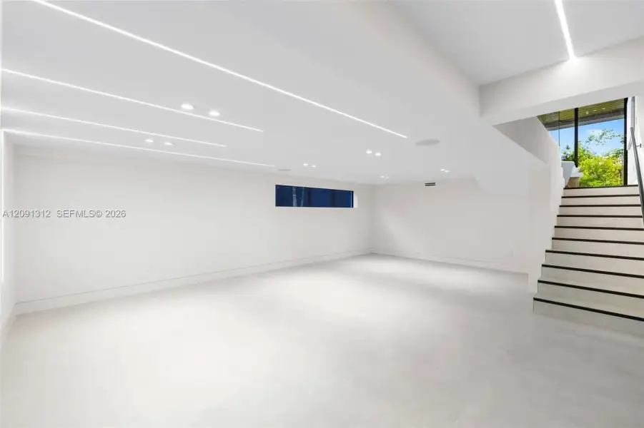Open floor plan featuring recessed linear lighting, smooth white walls, large-format tile flooring, and a staircase with a metal handrail Open floor plan featuring recessed linear lighting, smooth white walls, large-format tile flooring, and a staircase with a metal handrail