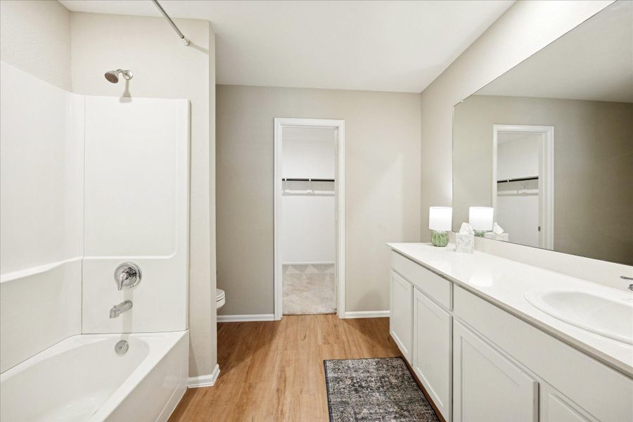 Bright and spacious Primary bathroom featuring a long vanity with mirror, a bathtub with a shower, and vinyl flooring. Connected to a walk-in closet for added convenience.