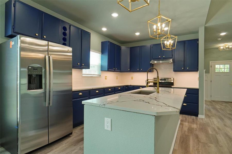 Kitchen featuring light stone countertops, light hardwood / wood-style flooring, blue cabinets, stainless steel fridge, and a kitchen island with sink