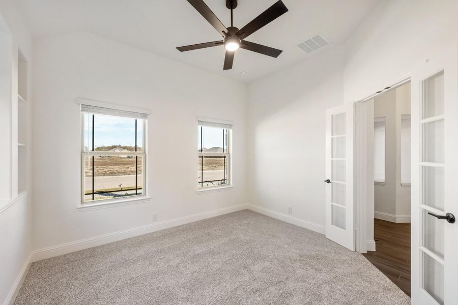 Empty bedroom with carpet, ceiling fan, two windows, and a French door leading to an ensuite bathroom Empty bedroom with carpet, ceiling fan, two windows, and a French door leading to an ensuite bathroom