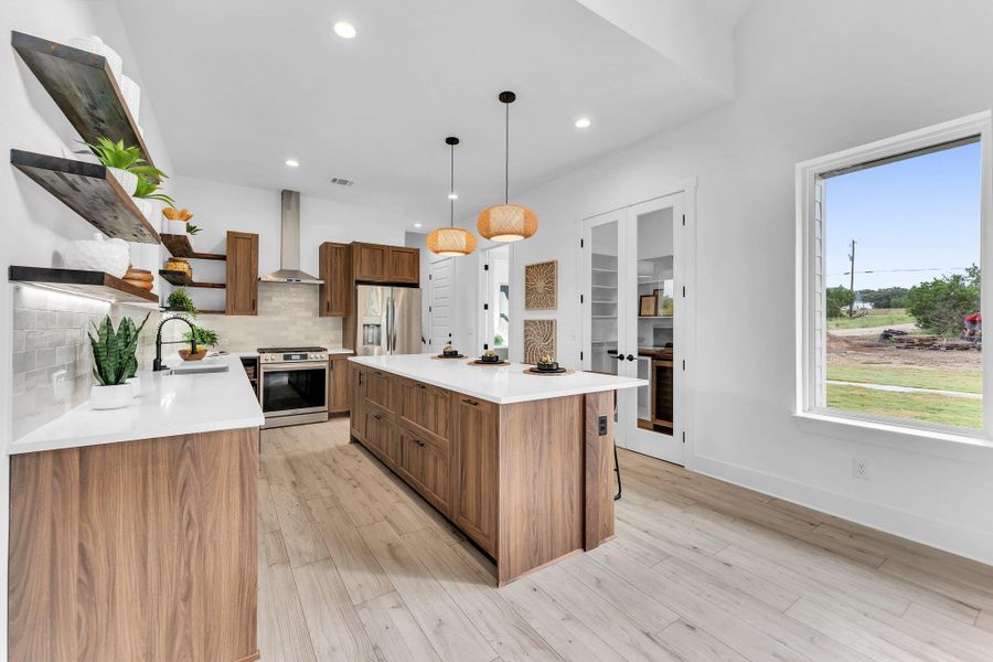 Kitchen featuring open shelves, pendant lighting, appliances with stainless steel finishes, brown cabinetry, and wall chimney range hood