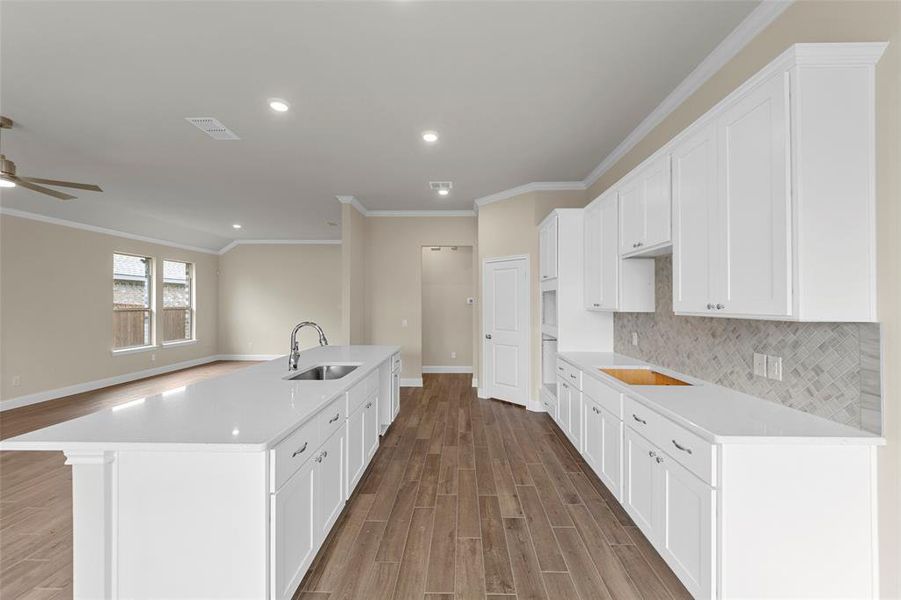 Modern kitchen featuring an extended island with a stainless steel sink, sleek white cabinetry, light-toned countertops, a diagonal tile backsplash, and recessed lighting