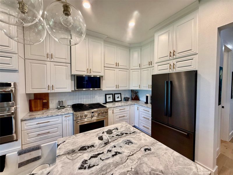 Gourmet kitchen featuring a substantial island with a patterned countertop, white shaker cabinetry, stainless steel oven and range, integrated microwave, and a black French door refrigerator - designer chandelier