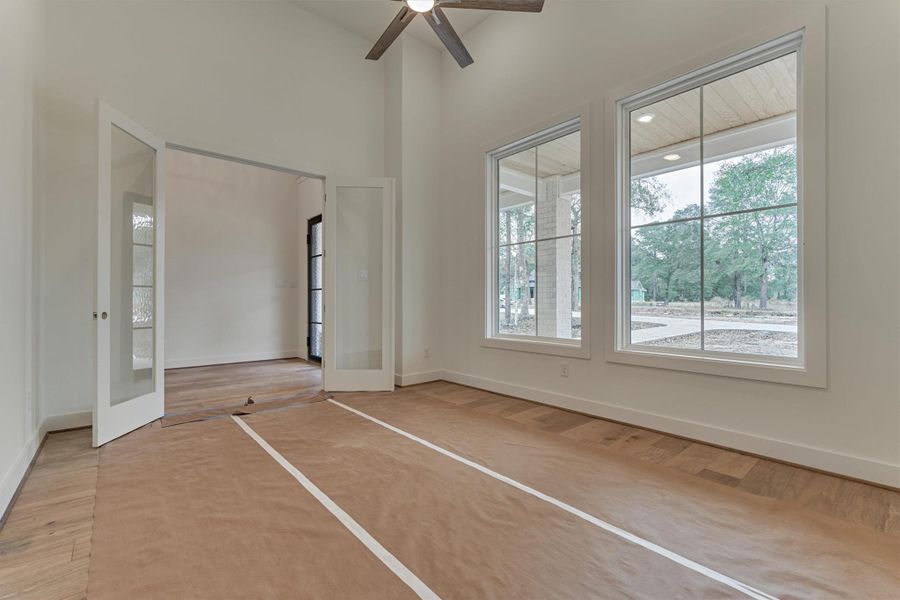 Spacious home office with glass French doors, tall ceilings, and large picture windows overlooking the peaceful front yard.