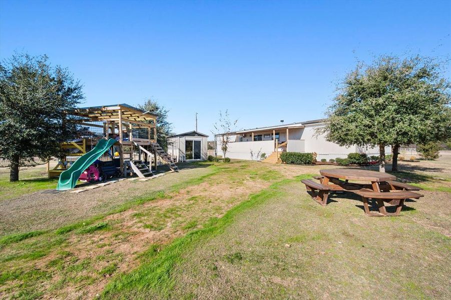 Expansive outdoor area featuring a wooden play structure with a green slide, a detached storage shed, a picnic table, and mature trees providing natural shade