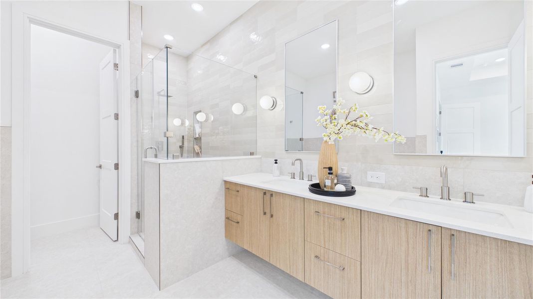 This bright and modern bathroom features a double vanity with sleek wooden cabinets, large mirrors, and contemporary fixtures. A spacious glass-enclosed shower complements the clean, minimalist design, enhanced by soft lighting and elegant tile work.