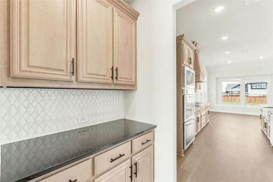 Kitchen with light wood cabinets, dark countertops, white tile backsplash, and built-in appliances on hardwood floors Kitchen with light wood cabinets, dark countertops, white tile backsplash, and built-in appliances on hardwood floors