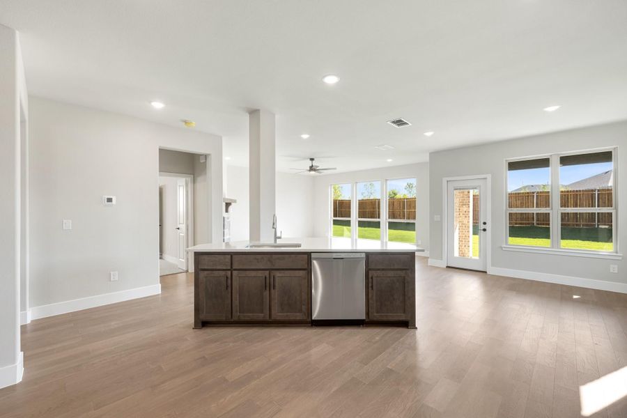Open-concept kitchen with dark wood island, stainless dishwasher, and hardwood floors overlooking bright living area with backyard windows.
