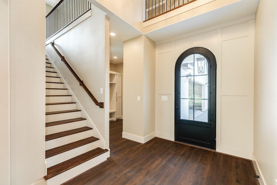 This elegant entryway features a dark wood floor and staircase, complemented by a striking black arched door with glass panes, creating a welcoming and stylish first impression.