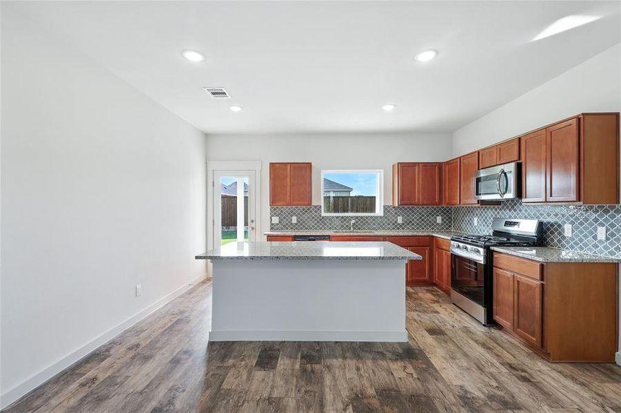 Kitchen with tasteful backsplash, appliances with stainless steel finishes, light stone counters, a center island, and dark wood-style flooring Kitchen with tasteful backsplash, appliances with stainless steel finishes, light stone counters, a center island, and dark wood-style flooring