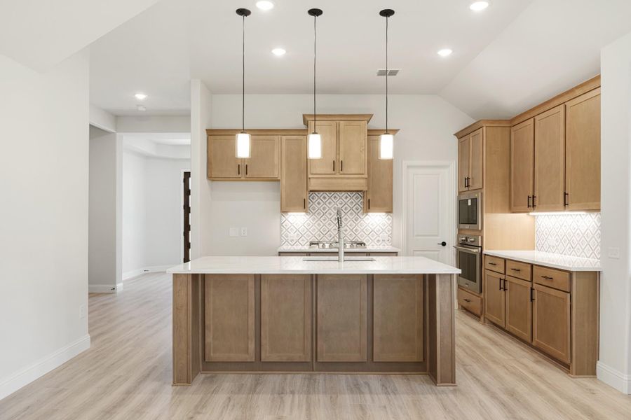 Kitchen with large center island, wood cabinets, patterned tile backsplash, and three pendant lights overhead.