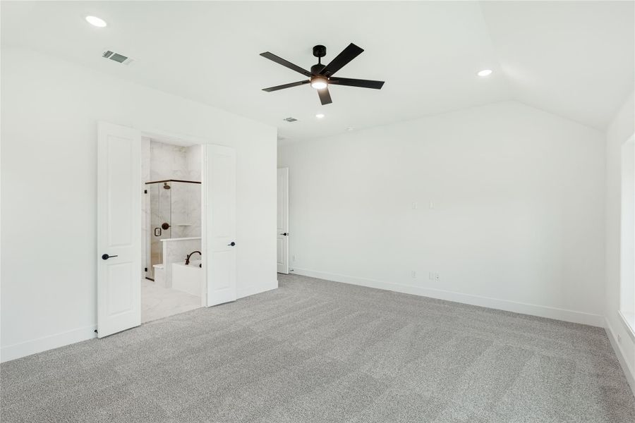 Spacious bedroom with gray carpet, ceiling fan, and open door to ensuite bathroom with soaking tub and shower Spacious bedroom with gray carpet, ceiling fan, and open door to ensuite bathroom with soaking tub and shower